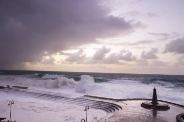 Büyük dalgaların fırtınası dalgakırana karşı kırılıyor ve Bajamar geziniyor gri fırtına bulutları ve arka planda bir deniz feneri ile.