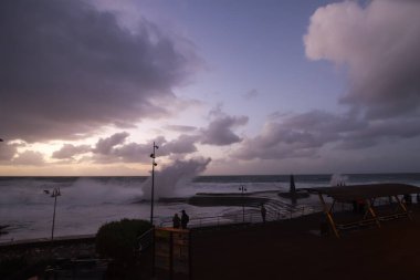 Büyük dalgaların fırtınası dalgakırana karşı kırılıyor ve Bajamar geziniyor gri fırtına bulutları ve arka planda bir deniz feneri ile.