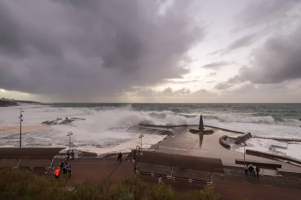 Büyük dalgaların fırtınası dalgakırana karşı kırılıyor ve Bajamar geziniyor gri fırtına bulutları ve arka planda bir deniz feneri ile.