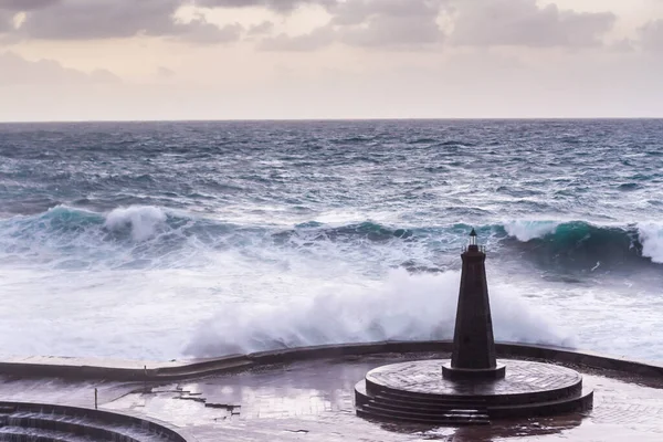 Büyük dalgaların fırtınası dalgakırana karşı kırılıyor ve Bajamar geziniyor gri fırtına bulutları ve arka planda bir deniz feneri ile.