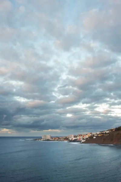 Günbatımı manzaralı okyanus manzarası ve kayalıklardan kayalık sahile vuran dalgalar ve arka planda altın bulutlar ve pitoresk bir köy. Bajamar, Tenerife, Kanarya Adaları. İspanya