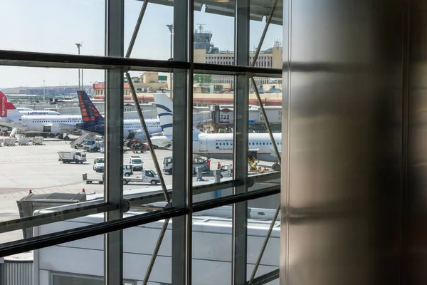 Yazın bir havaalanının yolcu terminali, yolcuların oturabileceği banklar, yürüyen bazı yolcular ve uçağın görülebileceği büyük pencereler..