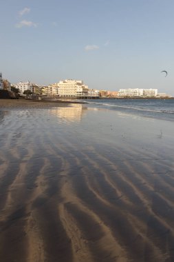 Gün batımında deniz manzarası, okyanus kıyısında ıslak sarı kumsal ve arka planda resimli bir balıkçı köyü. Playa Grande, El Medano, Tenerife, Kanarya Adaları. İspanya
