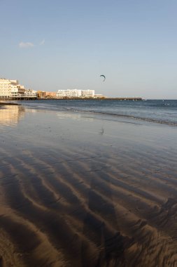 Gün batımında deniz manzarası, okyanus kıyısında ıslak sarı kumsal ve arka planda resimli bir balıkçı köyü. Playa Grande, El Medano, Tenerife, Kanarya Adaları. İspanya