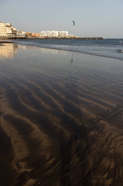 Gün batımında deniz manzarası, okyanus kıyısında ıslak sarı kumsal ve arka planda resimli bir balıkçı köyü. Playa Grande, El Medano, Tenerife, Kanarya Adaları. İspanya