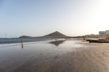 Günbatımında sarı kumlu sahil manzarası, okyanus sahildeki ıslak kumu yıkarken, güneş bulutsuz mavi gökyüzüyle arka plandaki dağları aydınlatıyor. Playa Grande, El Medano, Tenerife, Kanarya Adaları. İspanya