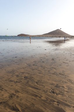 Günbatımında sarı kumlu sahil manzarası, okyanus sahildeki ıslak kumu yıkarken, güneş bulutsuz mavi gökyüzüyle arka plandaki dağları aydınlatıyor. Playa Grande, El Medano, Tenerife, Kanarya Adaları. İspanya