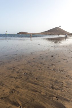 Günbatımında sarı kumlu sahil manzarası, okyanus sahildeki ıslak kumu yıkarken, güneş bulutsuz mavi gökyüzüyle arka plandaki dağları aydınlatıyor. Playa Grande, El Medano, Tenerife, Kanarya Adaları. İspanya