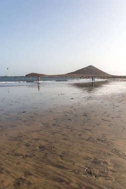 Günbatımında sarı kumlu sahil manzarası, okyanus sahildeki ıslak kumu yıkarken, güneş bulutsuz mavi gökyüzüyle arka plandaki dağları aydınlatıyor. Playa Grande, El Medano, Tenerife, Kanarya Adaları. İspanya