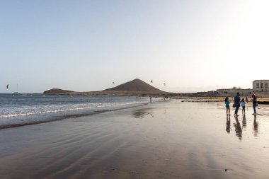 Günbatımında sarı kumlu sahil manzarası, okyanus sahildeki ıslak kumu yıkarken, güneş bulutsuz mavi gökyüzüyle arka plandaki dağları aydınlatıyor. Playa Grande, El Medano, Tenerife, Kanarya Adaları. İspanya