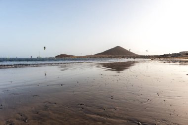 Günbatımında sarı kumlu sahil manzarası, okyanus sahildeki ıslak kumu yıkarken, güneş bulutsuz mavi gökyüzüyle arka plandaki dağları aydınlatıyor. Playa Grande, El Medano, Tenerife, Kanarya Adaları. İspanya