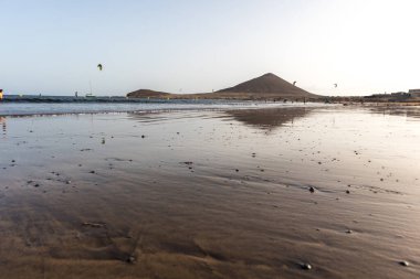 Günbatımında sarı kumlu sahil manzarası, okyanus sahildeki ıslak kumu yıkarken, güneş bulutsuz mavi gökyüzüyle arka plandaki dağları aydınlatıyor. Playa Grande, El Medano, Tenerife, Kanarya Adaları. İspanya