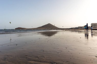 Günbatımında sarı kumlu sahil manzarası, okyanus sahildeki ıslak kumu yıkarken, güneş bulutsuz mavi gökyüzüyle arka plandaki dağları aydınlatıyor. Playa Grande, El Medano, Tenerife, Kanarya Adaları. İspanya