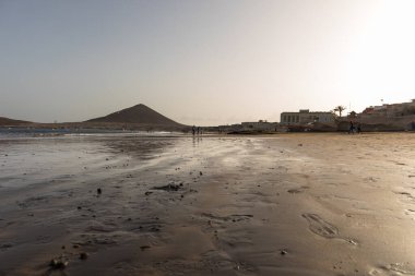 Günbatımında sarı kumlu sahil manzarası, okyanus sahildeki ıslak kumu yıkarken, güneş bulutsuz mavi gökyüzüyle arka plandaki dağları aydınlatıyor. Playa Grande, El Medano, Tenerife, Kanarya Adaları. İspanya