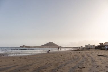 Günbatımında sarı kumlu sahil manzarası, okyanus sahildeki ıslak kumu yıkarken, güneş bulutsuz mavi gökyüzüyle arka plandaki dağları aydınlatıyor. Playa Grande, El Medano, Tenerife, Kanarya Adaları. İspanya