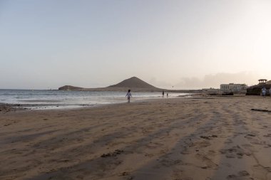Günbatımında sarı kumlu sahil manzarası, okyanus sahildeki ıslak kumu yıkarken, güneş bulutsuz mavi gökyüzüyle arka plandaki dağları aydınlatıyor. Playa Grande, El Medano, Tenerife, Kanarya Adaları. İspanya