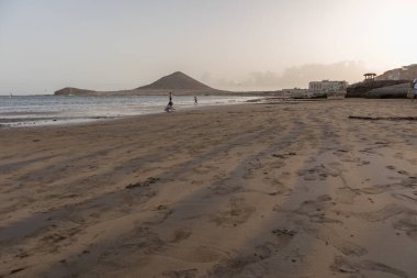 Günbatımında sarı kumlu sahil manzarası, okyanus sahildeki ıslak kumu yıkarken, güneş bulutsuz mavi gökyüzüyle arka plandaki dağları aydınlatıyor. Playa Grande, El Medano, Tenerife, Kanarya Adaları. İspanya