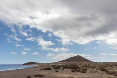 Günbatımında sarı kumlu sahil manzarası, okyanus sahildeki ıslak kumu yıkarken, güneş de büyük beyaz bulutlu mavi gökyüzü ile arka plandaki dağları aydınlatır. Playa Grande, El Medano, Tenerife, Kanarya Adaları. İspanya