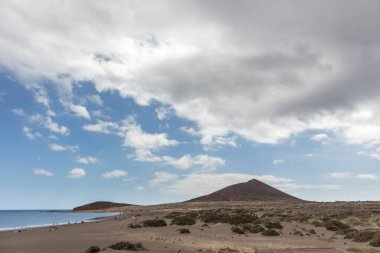Günbatımında sarı kumlu sahil manzarası, okyanus sahildeki ıslak kumu yıkarken, güneş de büyük beyaz bulutlu mavi gökyüzü ile arka plandaki dağları aydınlatır. Playa Grande, El Medano, Tenerife, Kanarya Adaları. İspanya