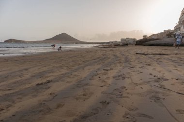 Günbatımında sarı kumlu sahil manzarası, okyanus sahildeki ıslak kumu yıkarken, güneş bulutsuz mavi gökyüzüyle arka plandaki dağları aydınlatıyor. Playa Grande, El Medano, Tenerife, Kanarya Adaları. İspanya