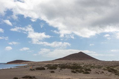 Günbatımında sarı kumlu sahil manzarası, okyanus sahildeki ıslak kumu yıkarken, güneş de büyük beyaz bulutlu mavi gökyüzü ile arka plandaki dağları aydınlatır. Playa Grande, El Medano, Tenerife, Kanarya Adaları. İspanya