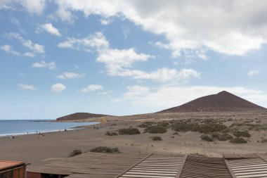 Günbatımında sarı kumlu sahil manzarası, okyanus sahildeki ıslak kumu yıkarken, güneş de büyük beyaz bulutlu mavi gökyüzü ile arka plandaki dağları aydınlatır. Playa Grande, El Medano, Tenerife, Kanarya Adaları. İspanya