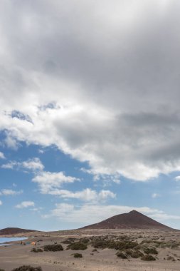 Günbatımında sarı kumlu sahil manzarası, okyanus sahildeki ıslak kumu yıkarken, güneş de büyük beyaz bulutlu mavi gökyüzü ile arka plandaki dağları aydınlatır. Playa Grande, El Medano, Tenerife, Kanarya Adaları. İspanya
