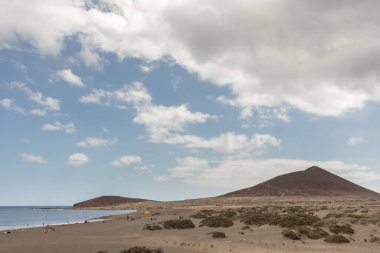 Günbatımında sarı kumlu sahil manzarası, okyanus sahildeki ıslak kumu yıkarken, güneş de büyük beyaz bulutlu mavi gökyüzü ile arka plandaki dağları aydınlatır. Playa Grande, El Medano, Tenerife, Kanarya Adaları. İspanya