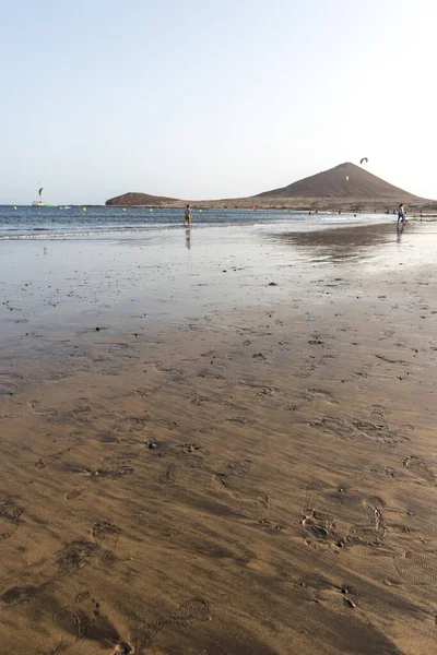 Günbatımında sarı kumlu sahil manzarası, okyanus sahildeki ıslak kumu yıkarken, güneş bulutsuz mavi gökyüzüyle arka plandaki dağları aydınlatıyor. Playa Grande, El Medano, Tenerife, Kanarya Adaları. İspanya