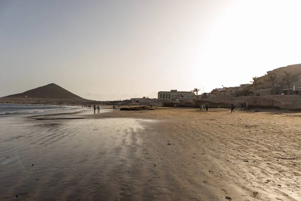 Günbatımında sarı kumlu sahil manzarası, okyanus sahildeki ıslak kumu yıkarken, güneş bulutsuz mavi gökyüzüyle arka plandaki dağları aydınlatıyor. Playa Grande, El Medano, Tenerife, Kanarya Adaları. İspanya