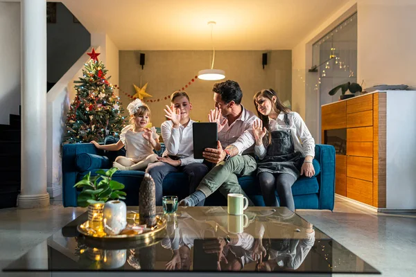 A father with his children sitting in the living room on the couch while they make a video call via tablet and show off their decorated Christmas tree. The room is decorated in a festive spirit, as well as the coming Christmas.