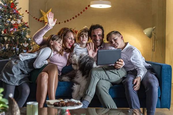 A happy family is sitting on the couch in living room and having a video call via tablet. The room is decorated in the Christmas spirit. Also, their dog is on the couch with them.