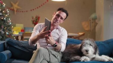 Adam oturma odasındaki kanepede oturuyor Noel süsleriyle, köpeğini okşuyor ve tablet üzerinde çalışıyor..