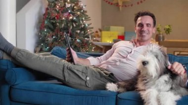 Adam oturma odasındaki kanepede oturuyor Noel süsleriyle, köpeğini okşuyor ve tablet üzerinde çalışıyor..