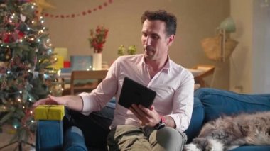 Köpeği yanında otururken adam kanepede oturuyor. Oturma odası Noel ruhuyla süslenmiş ve tabletiyle birine Noel hediyesi gösteren bir video görüşmesi yapıyor..