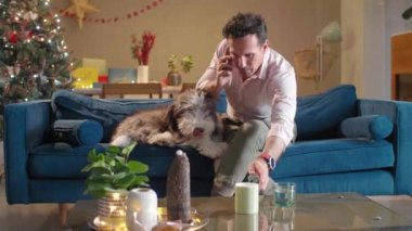 Adam telefonda konuşurken kanepede oturuyor. Aynı zamanda, o da kanepede yatan köpeğini okşar. Oturma odası Noel ruhuyla süslenmiş..