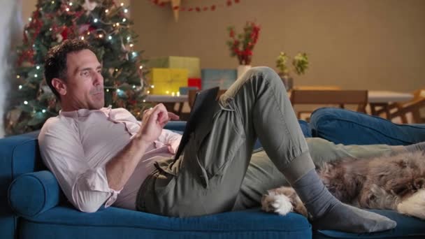 Чоловік Сидить Дивані Вітальні Різдвяними Прикрасами Гладить Собаку Працює Планшеті — стокове відео