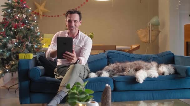 Чоловік Сидить Дивані Його Собака Сидить Поруч Вітальня Прикрашена Різдвяним — стокове відео