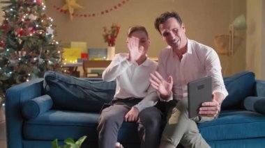 Bir baba ve oğlu oturma odasında kanepede otururken onlar tabletle video görüşmesi yapıp süslü Noel ağaçlarıyla hava atıyorlar. Oda Noel ruhuyla süslenmiş..