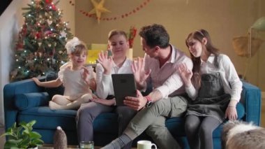 Çocuklarıyla birlikte oturma odasında kanepede oturan bir baba onlar tabletle video görüşmesi yaparken ve süslü Noel ağaçlarıyla hava atarken. Oda şenlikli bir ruhla dekore edildi. Gelecek Noel 'le birlikte..