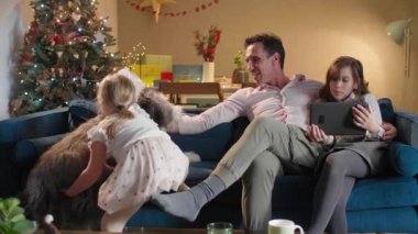 Bir baba, çocuklarının oturma odasında kanepede otururken onlar tabletten bir şey izliyorlar. Oda şenlikli bir ruhla dekore edildi. Gelecek Noel 'le birlikte..