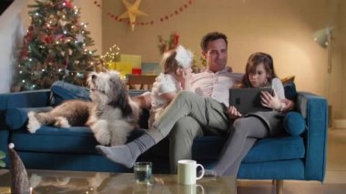 Adam oturma odasındaki kanepede iki kızı ve köpeğiyle oturuyor. Oda, yaklaşan bayram ve Noel ruhuyla dekore edildi. Tablette bir şey arıyorlar..