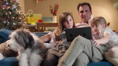 Adam oturma odasındaki kanepede iki kızı ve köpeğiyle oturuyor. Oda, yaklaşan bayram ve Noel ruhuyla dekore edildi. Tablette bir şey arıyorlar..