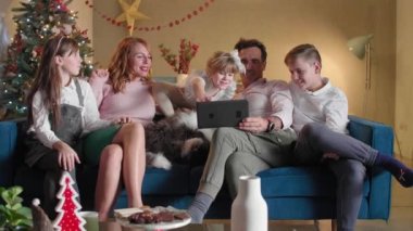 Mutlu bir aile oturma odasındaki kanepede oturuyor ve tabletle video görüşmesi yapıyor. Oda Noel ruhuyla süslenmiş. Ayrıca, köpekleri de onlarla birlikte kanepede..