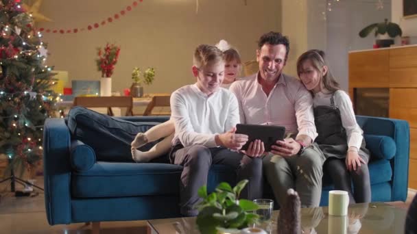 A father sits with his children in the living room on the couch while they watching something on a tablet. The room is decorated in a festive spirit, as well as the coming Christmas. — Stock Footage Father Sits His Children Living Room Couch While Watching Something Stock Video