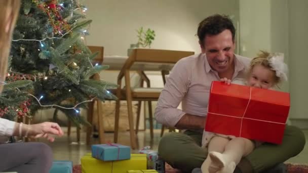 父は贈り物を交換している間 娘たちと一緒にクリスマスツリーの隣の床に座っています 雰囲気はお祝いで 部屋は休日の精神で飾られています — ストック動画
