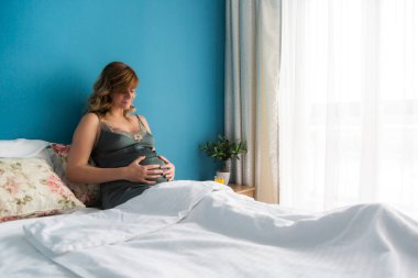 Yarı-oturma pozisyonunda, elleri karnında gülümseyen hamile bir kadın. Karnına doğru bakıyor..