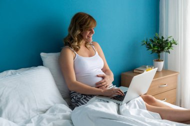 Dizüstü bilgisayarı üzerinde çalışırken yatağında oturan gülümseyen ve mutlu, hamile, beyaz bir kadın. Bir eli karnında..
