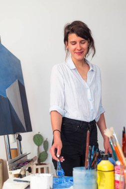 Stüdyosunda, gülümseyen genç bir ressam palette fırçayla boya sürüyor. Etrafında bir sehpaya yerleştirilmiş sanat eserinin yanı sıra resim yapmak için diğer malzemeler yer alıyor..