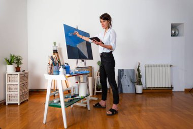Genç ressam onun stüdyosunda duruyor ve tabletten eskizi doğrudan tablonun üzerine kopyalıyor. Arka planda resim yapmak için gerekli malzemeyi görebilirsiniz..